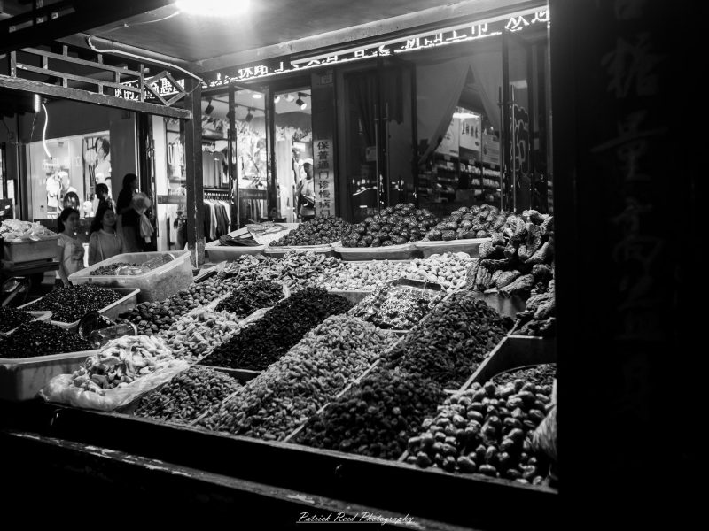 Black-and-white photograph of a market stall displaying rows of dried fruit and nuts beneath illuminated shop signs.