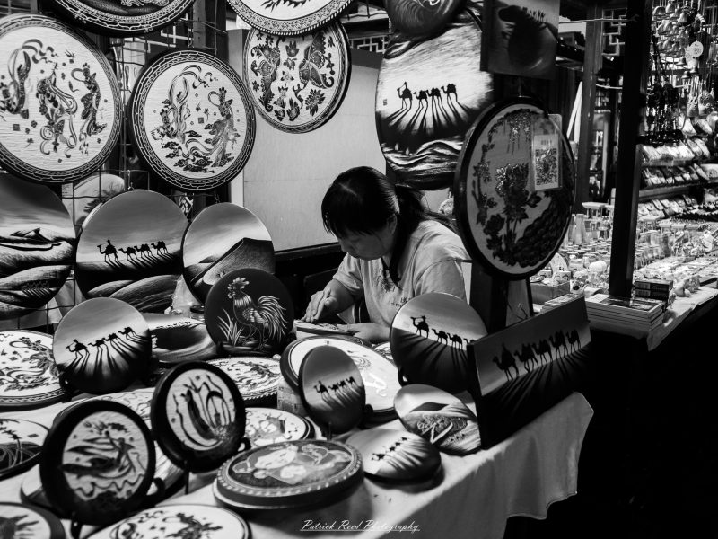 Black-and-white photograph of a craftsperson working behind a market table covered with decorative plates showing camels, birds and figures.