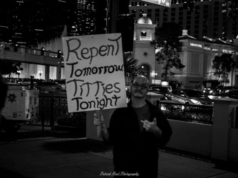 A man holds a handwritten “Repent Tomorrow, Titties Tonight” sign on a Las Vegas sidewalk at night, photographed in black and white.