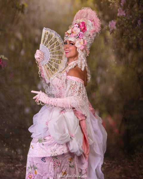 Costumed woman in pink holding an ornate lace fan, surrounded by softly blurred spring flowers.