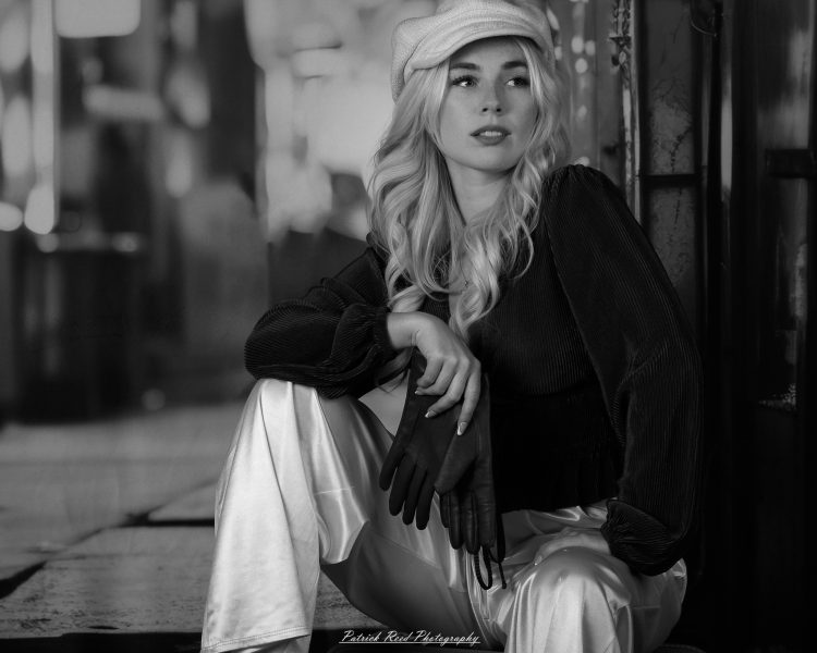 Black-and-white portrait of a woman in a newsboy cap seated with one knee raised and black gloves draped over it.