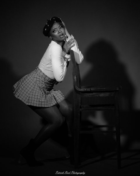 Black-and-white studio portrait of a woman in a beret and checked skirt leaning against a tall wooden chair, hands clasped near her face.