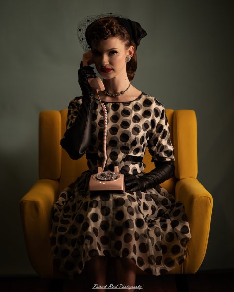 Retro studio portrait of a woman in a polka-dot dress holding a pink rotary telephone while seated in a yellow armchair.
