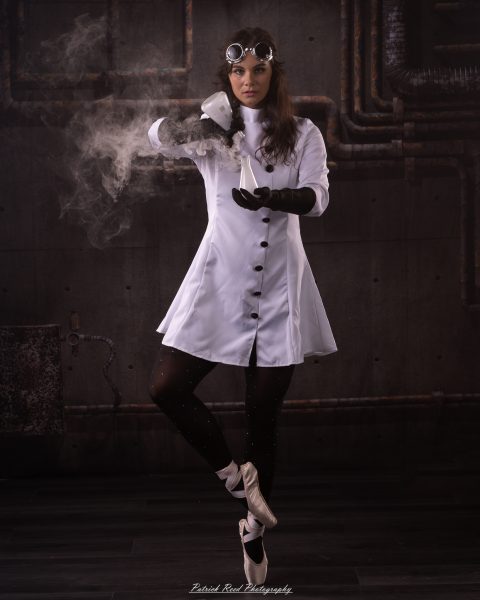 Woman in a white lab coat and goggles holds a flask, releasing steam in an industrial setting.