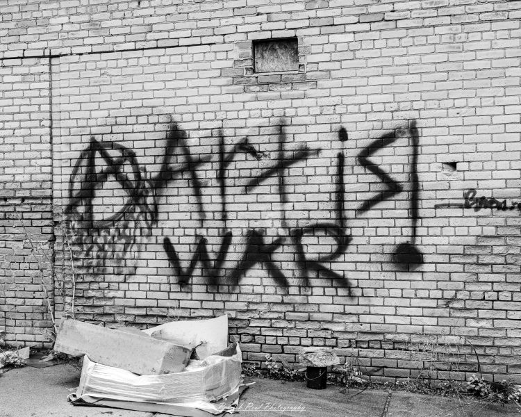 Black-and-white photograph of “ART IS WAR!” graffiti on a brick wall above discarded packaging.