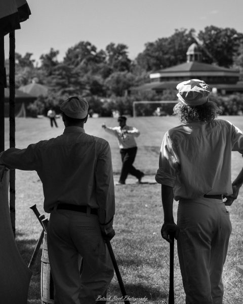 Reenactors in period clothing stand with rifles outdoors, watching a grassy field and a historic building.