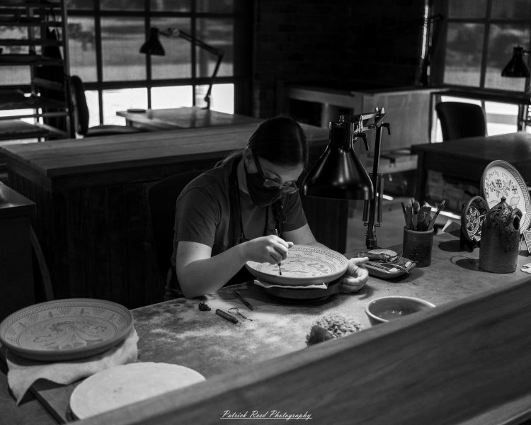 Black-and-white photograph of a masked craftsperson decorating a ceramic plate at a lamp-lit workbench in Greenfield Village.