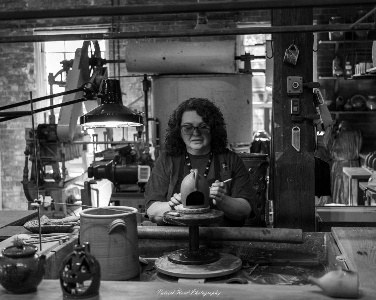 Black-and-white portrait of a craftsperson shaping a small ceramic structure at a workbench surrounded by pottery in Greenfield Village.