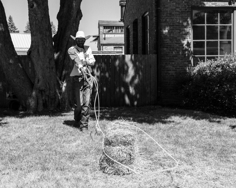 A man in a cowboy hat loops a lasso around a hay bale at Greenfield Village, photographed in black and white.