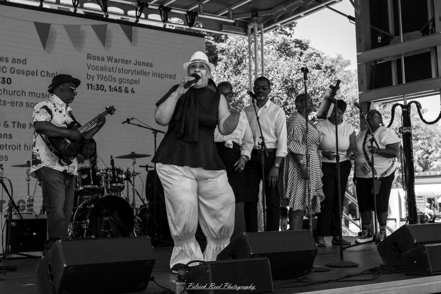 Black-and-white photograph of a singer in a light hat performing with a band and backing vocalists on an outdoor stage at Greenfield Village.