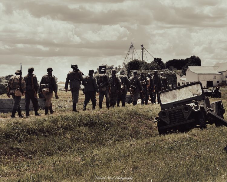 Vintage military reenactors march in formation past a parked jeep on an open field.