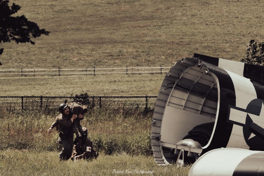 Reenactors crouch beside a vintage military aircraft in a grassy field during a historical demonstration.
