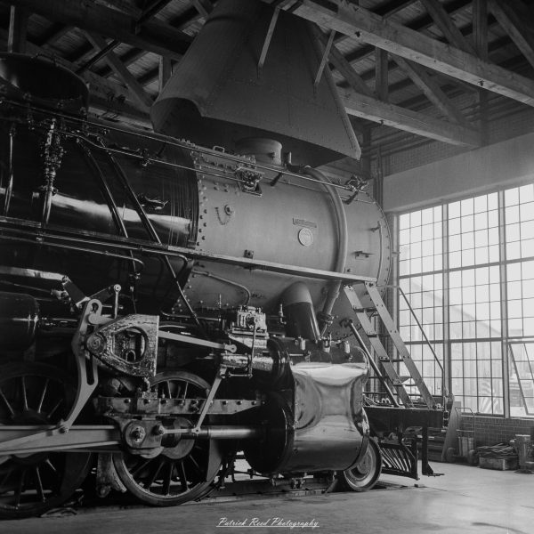 Close-up of a steam locomotive’s front and wheel assembly inside an industrial train hall.