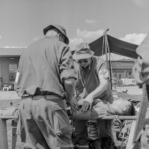 Attendants in vintage clothing tend to a patient lying on a stretcher inside a field tent.