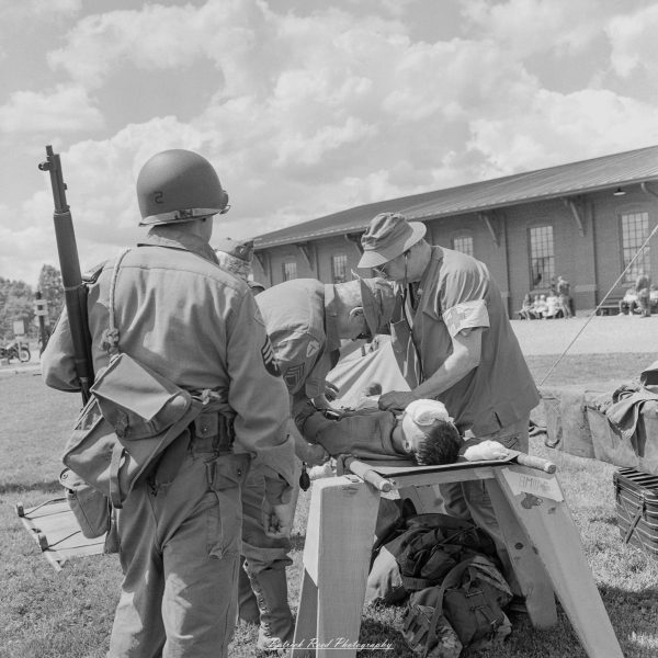 Historical reenactors in uniforms aid a soldier lying on a stretcher near a large event tent.