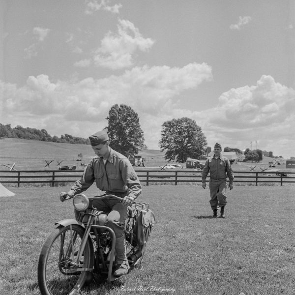 A reenactor rides a vintage motorcycle across a grassy field while another participant walks nearby.