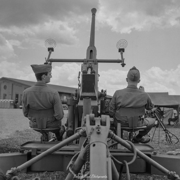 Two men in vintage uniforms stand at a military cannon during a historical reenactment in an open field.