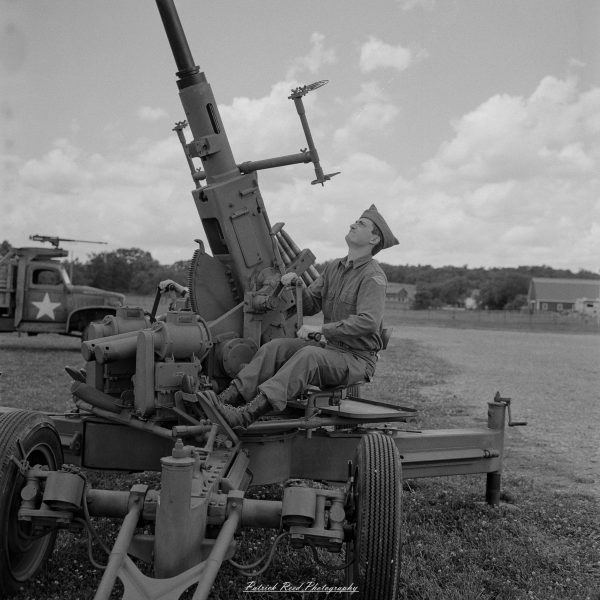 Soldier seated beside a vintage steam-powered artillery gun, outdoors under a cloudy sky at an event.
