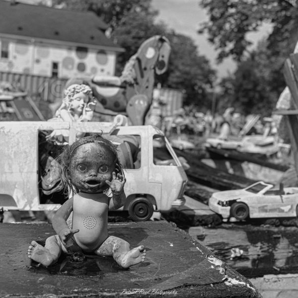 Black-and-white close-up of a weathered doll with one hand raised, surrounded by toy vehicles in an outdoor art display.