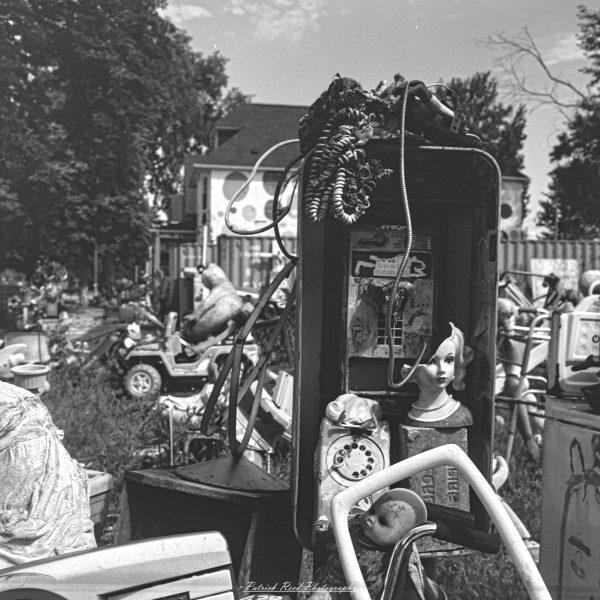 Black-and-white photograph of an outdoor art assemblage combining an old telephone housing, rotary phone, cords and a mannequin bust.