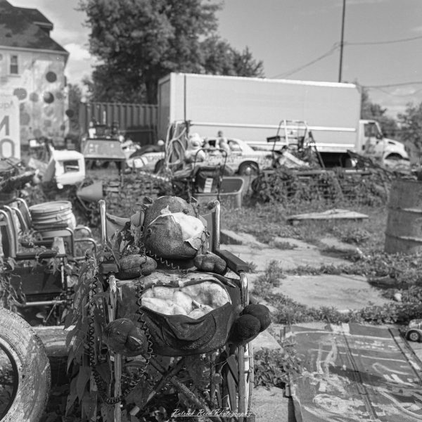 Black-and-white photograph of a torn stuffed toy seated in a weathered wheelchair amid an outdoor found-object art installation.