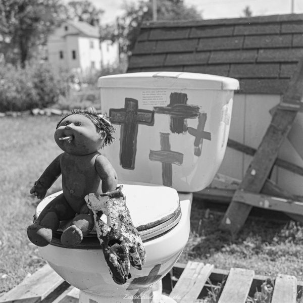 Black-and-white photograph of a weathered doll seated on a discarded toilet painted with crosses in an outdoor art display.