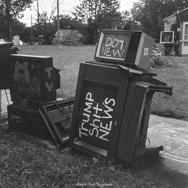 Black-and-white photograph of discarded televisions painted with messages about news and politics in an outdoor art installation.