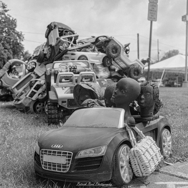 Black-and-white photograph of mannequin heads in a child-sized Audi convertible, with a pile of toy vehicles behind.
