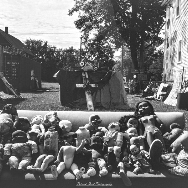 Black-and-white photograph of a couch crowded with weathered dolls in an outdoor yard art installation.