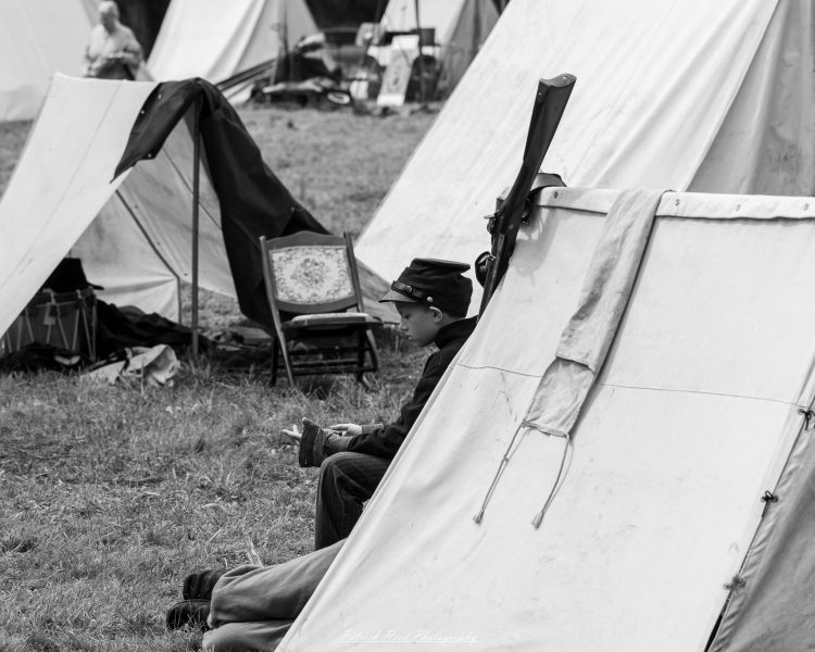 Civil War reenactor seated in a white canvas camp tent, holding a rifle during a historical event.