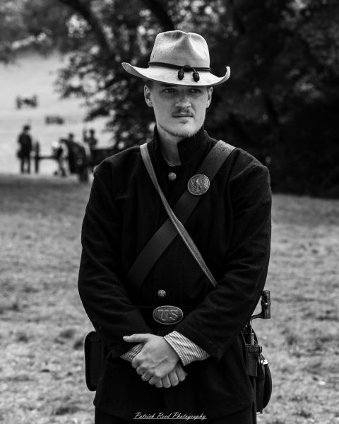 Civil War reenactor in dark uniform and slouch hat stands outdoors with hands clasped during event.