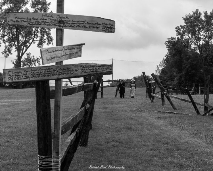 Civil War reenactment yard with handwritten wooden signs and two period-dressed figures walking in grass.