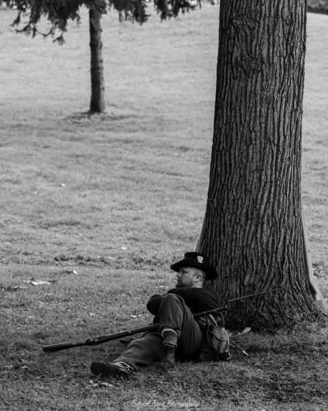 Civil War reenactor resting beside a tree, holding a rifle in an outdoor black-and-white scene.