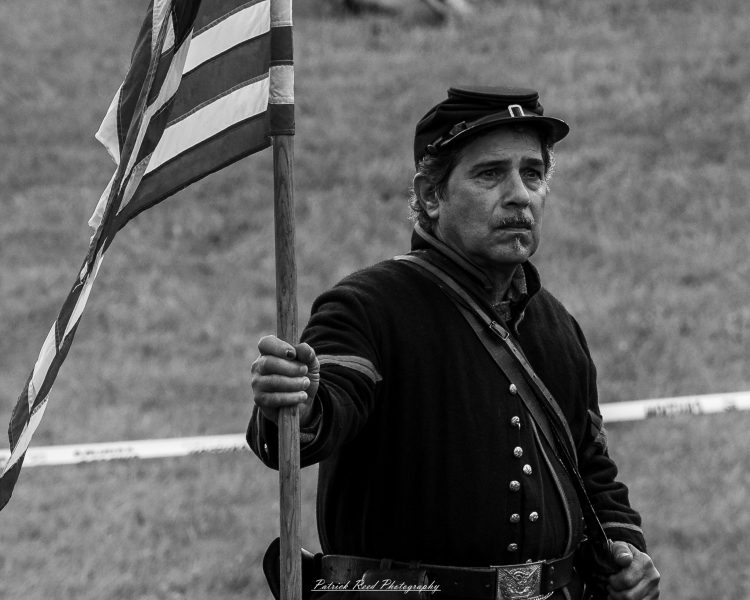 Civil War reenactor stands in a field holding a striped Union flag during a historical event.