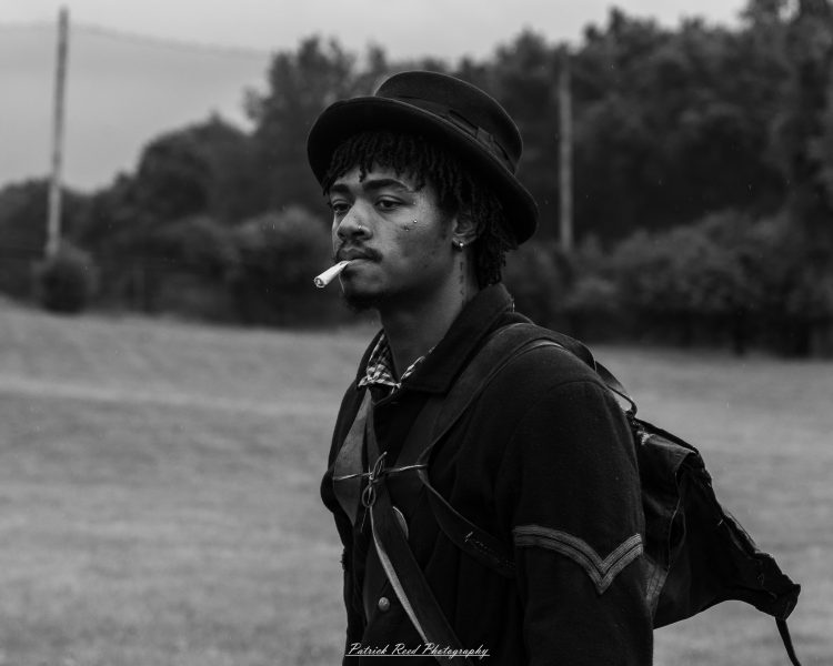 Civil War reenactor in period hat smoking a cigarette outdoors near trees and open field