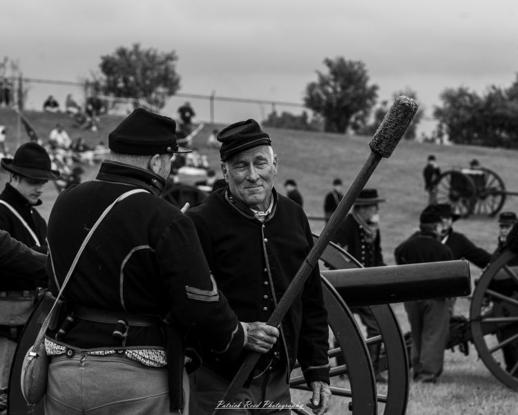 Civil War reenactors in period uniforms stand beside an antique cannon at an outdoor battlefield event.