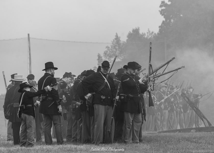 Civil War reenactors in dark uniforms stand in formation with rifles amid smoke and morning haze.