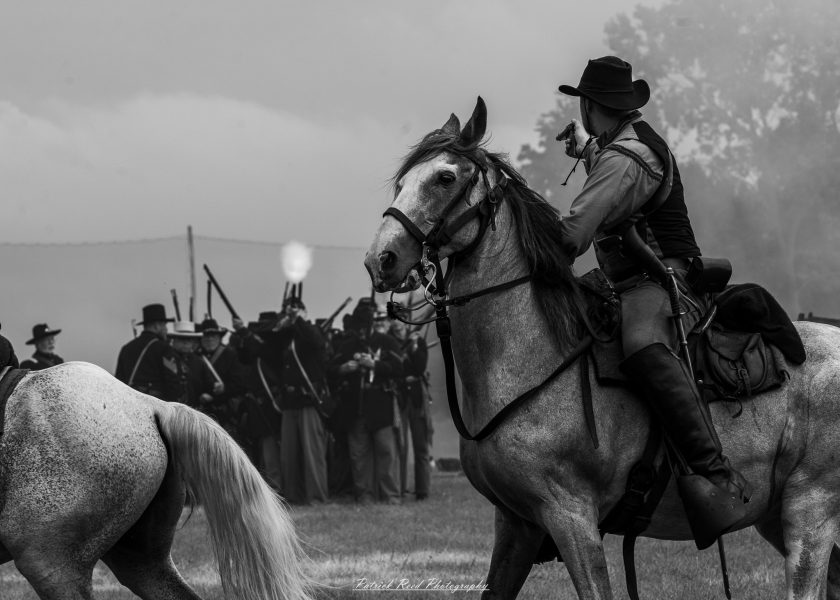 Civil War reenactor in a hat sits on a horse as mounted cavalry watches in a field.