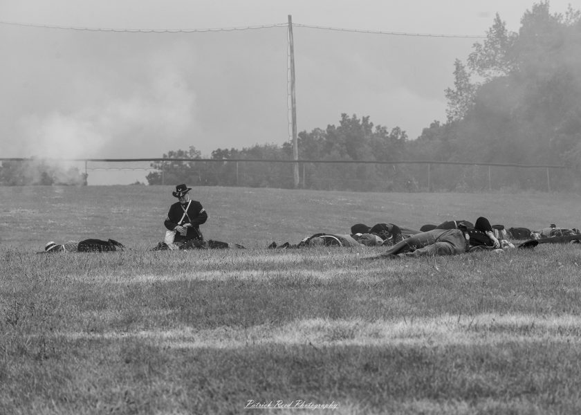 Civil War reenactment soldier kneels beside a line of troops in a grassy field under foggy sky.