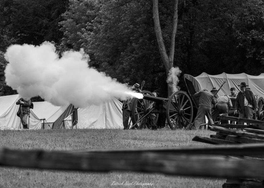 Civil War reenactors fire a cannon with thick smoke near a canvas camp tent in a tree-lined field.