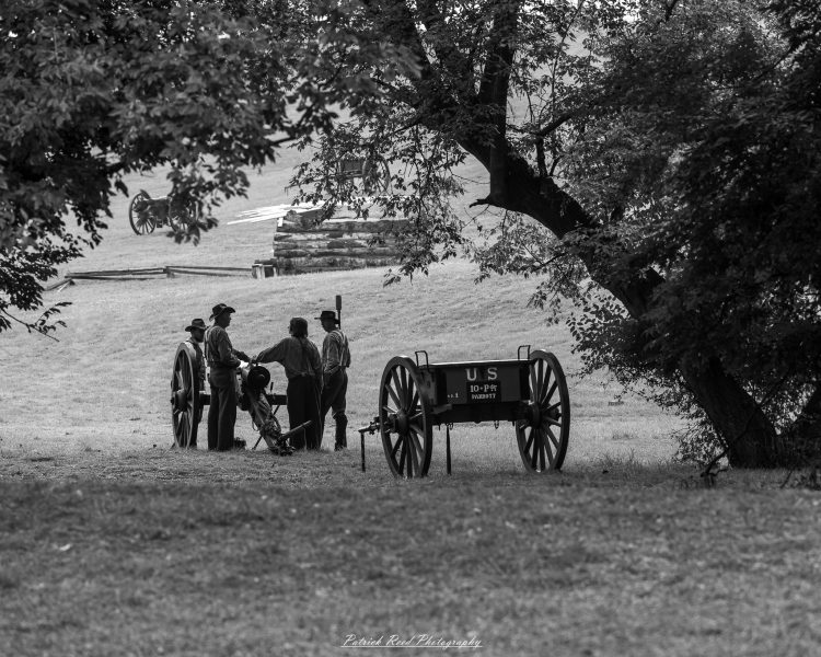 Civil War reenactors gather beside a mounted Parrott cannon in a tree-lined field.