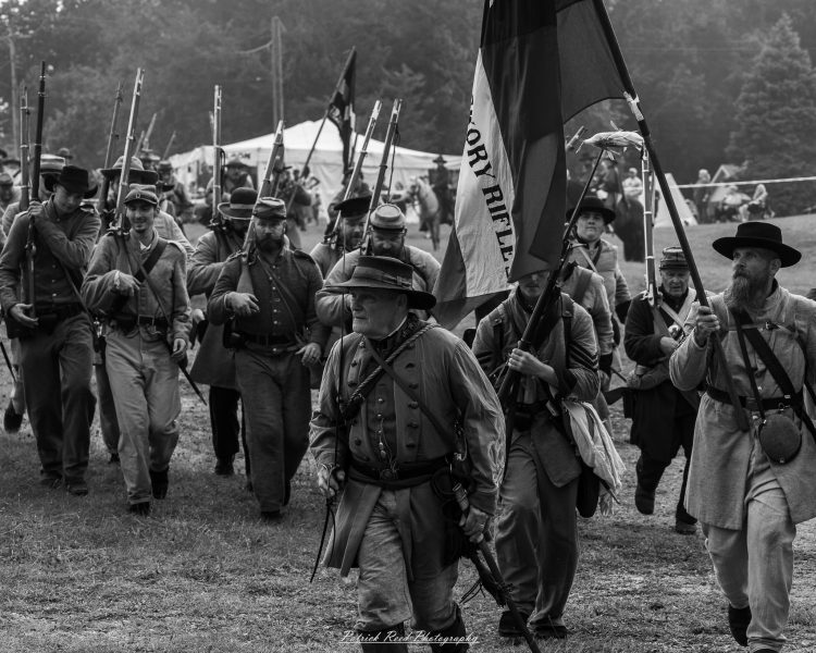 Civil War reenactors march with rifles and a banner through a wooded camp during an outdoor event.