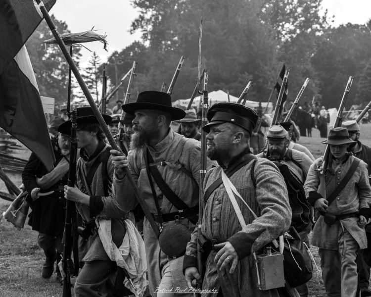 Civil War reenactors march in black-and-white, holding rifles and waving flags outdoors among spectators.