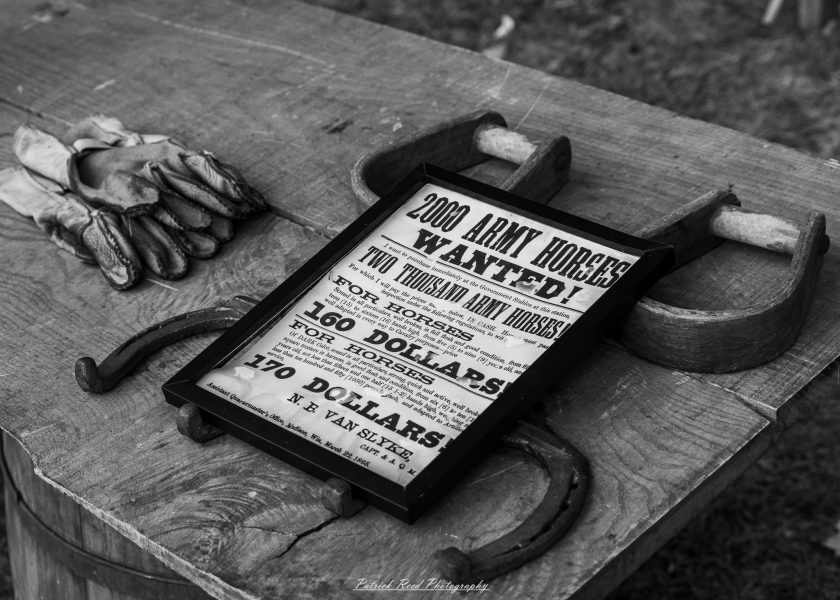Newspaper clipping reading “2000 Army Homes” lies on a wooden barrel beside folded gloves.