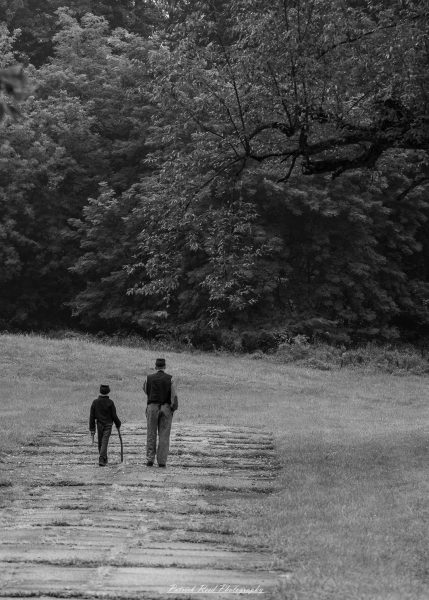 Two Civil War reenactors in period clothing walk along a dirt path through grass with trees behind them.