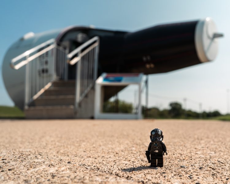 Toy pilot figure standing on runway gravel, with a military jet in the background under daylight.