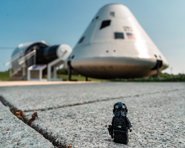 Toy soldier figure kneeling on gravel near parked transport plane on runway under clear blue sky.