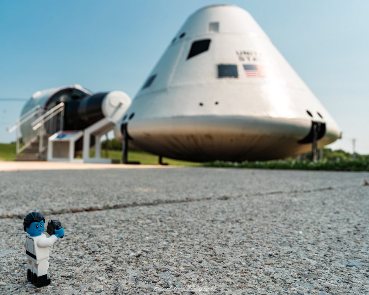Lego minifigure in foreground with a military jet on display under a clear blue sky.