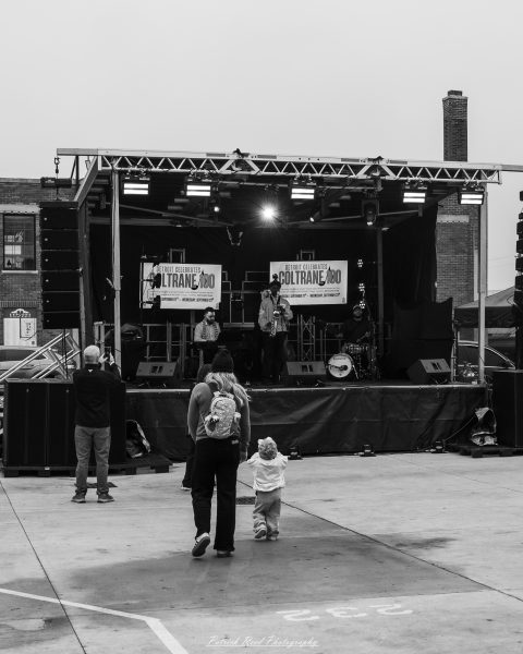 An adult and child approach an outdoor jazz stage beneath Detroit Celebrates Coltrane 100 banners at Eastern Market.