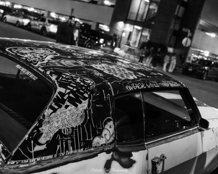 Graffiti-covered classic car at night with blurred market lights and pedestrians in the background.
